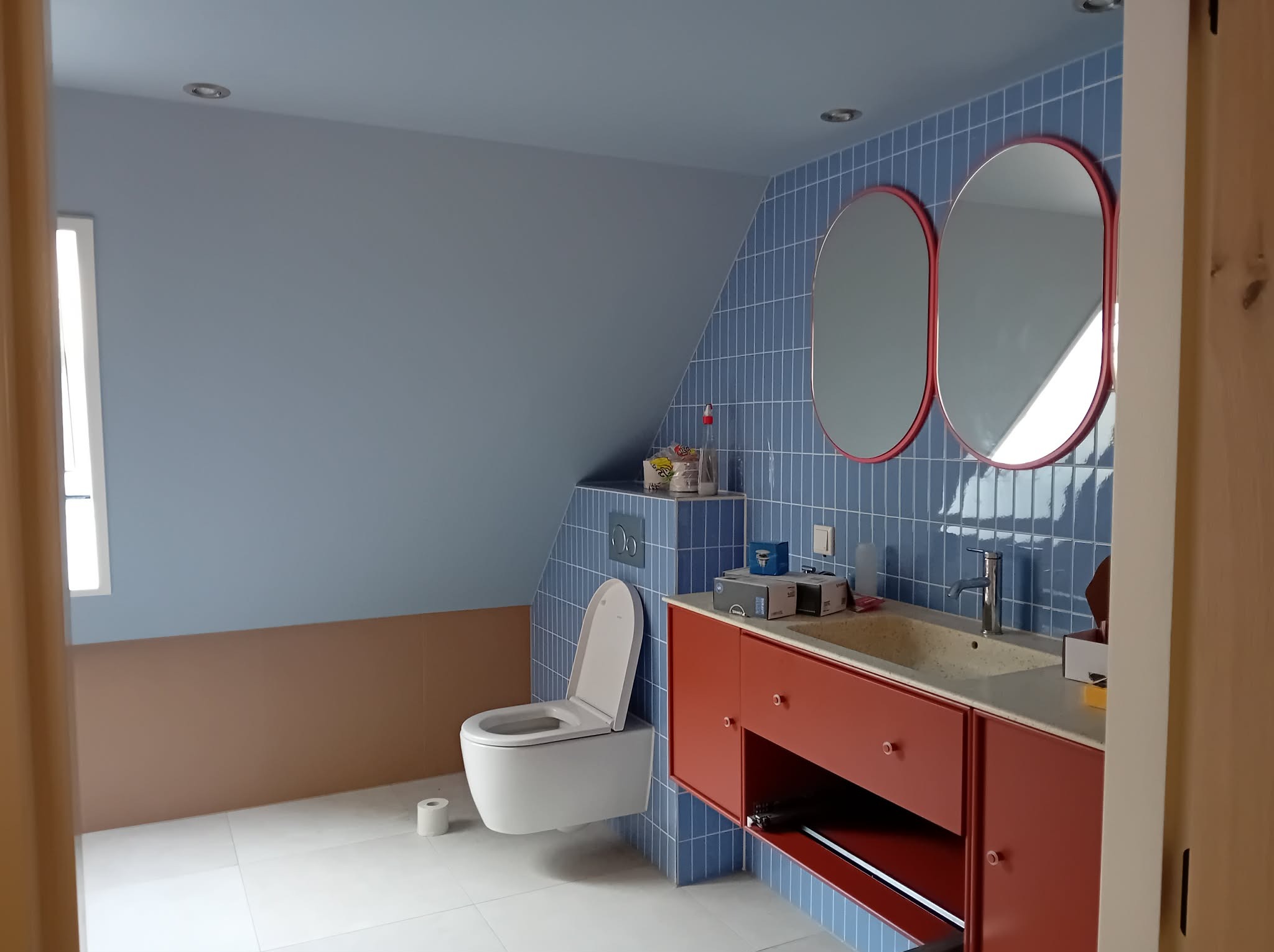Niebieska łazienka na poddaszu z owalnymi lustrami w czerwonych ramach, wiszącą toaletą i czerwoną szafką pod umywalką. Ukończony remont, widok zza framugi.