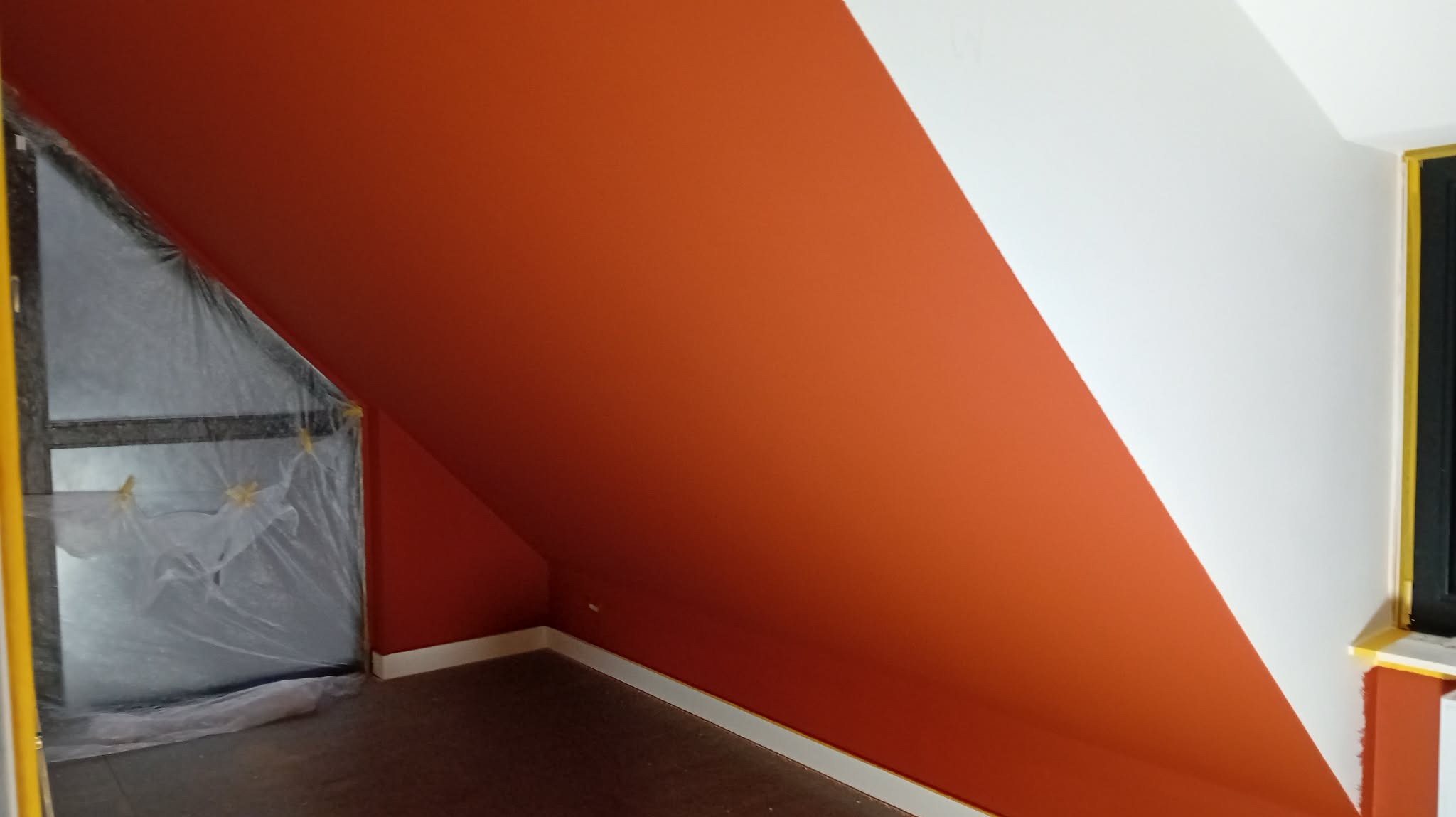 Pomarańczowo-białe malowanie skośnej ściany poddasza, okno zabezpieczone folią malarską, widoczne taśmy malarskie, podłoga zabezpieczona.