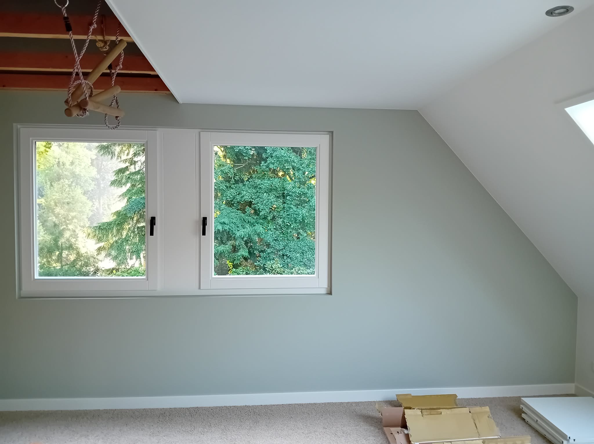 Pokój na poddaszu po malowaniu ścian na jasny, pastelowy kolor. Widoczne okna z widokiem na zieleń, drewniane elementy konstrukcyjne sufitu i kartony na podłodze.