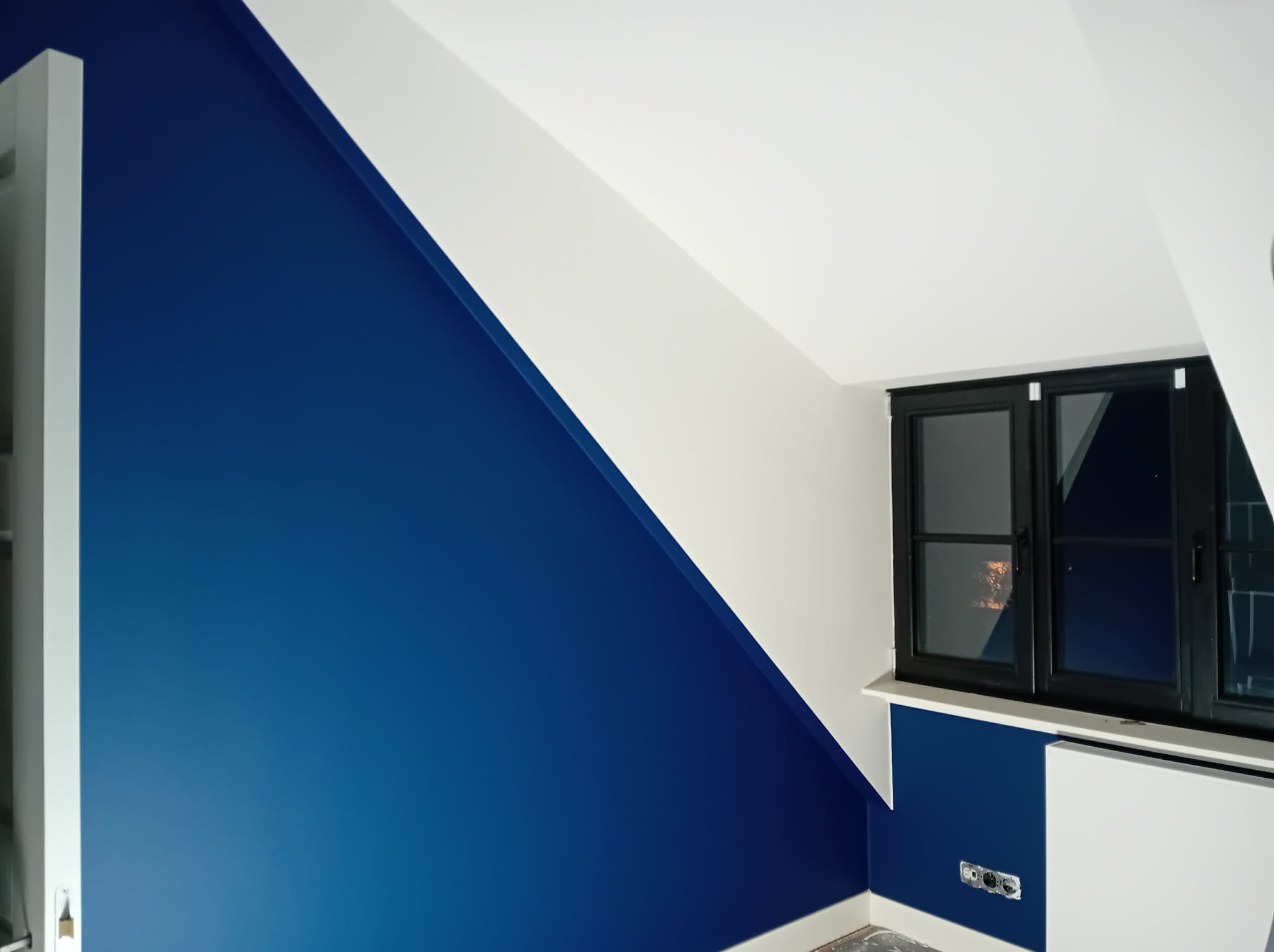 Ściana w odcieniach bieli i intensywnego granatu w pokoju na poddaszu z oknem w czarnej ramie. Widoczne gniazdka elektryczne i fragment grzejnika.