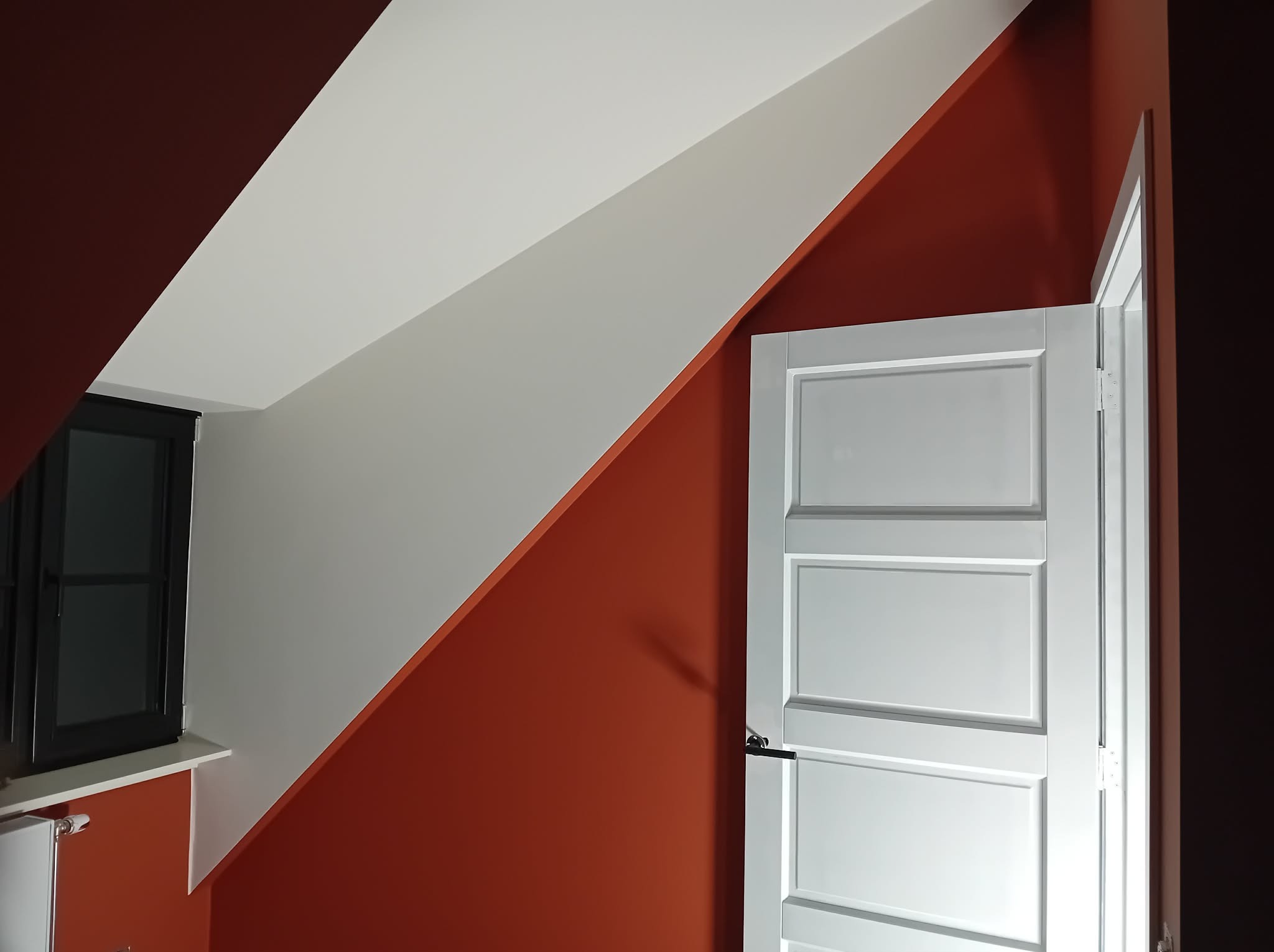 Pomarańczowa ściana w pokoju ze skośnym sufitem, białe wykończenie, czarne okno i białe drzwi z trzema panelami. Świeżo po malowaniu.