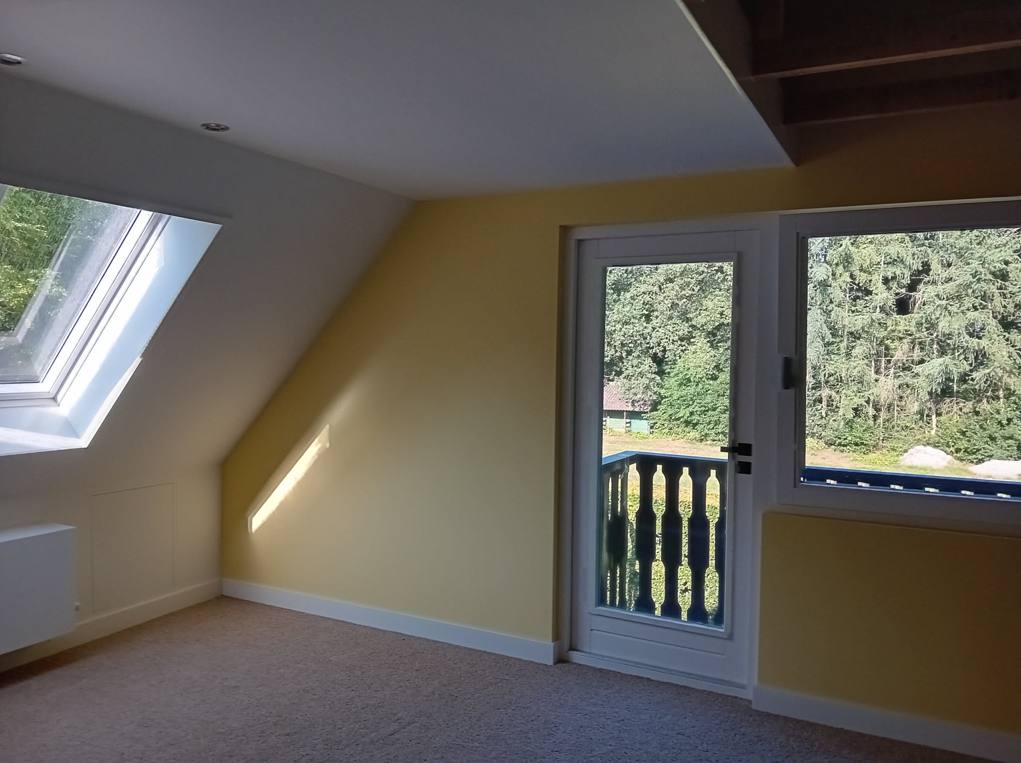 Żółty pokój na poddaszu z oknem dachowym i wyjściem na balkon z widokiem na zieleń. Ściany pomalowane na jasnożółto, białe listwy przypodłogowe i wykładzina dywanowa.