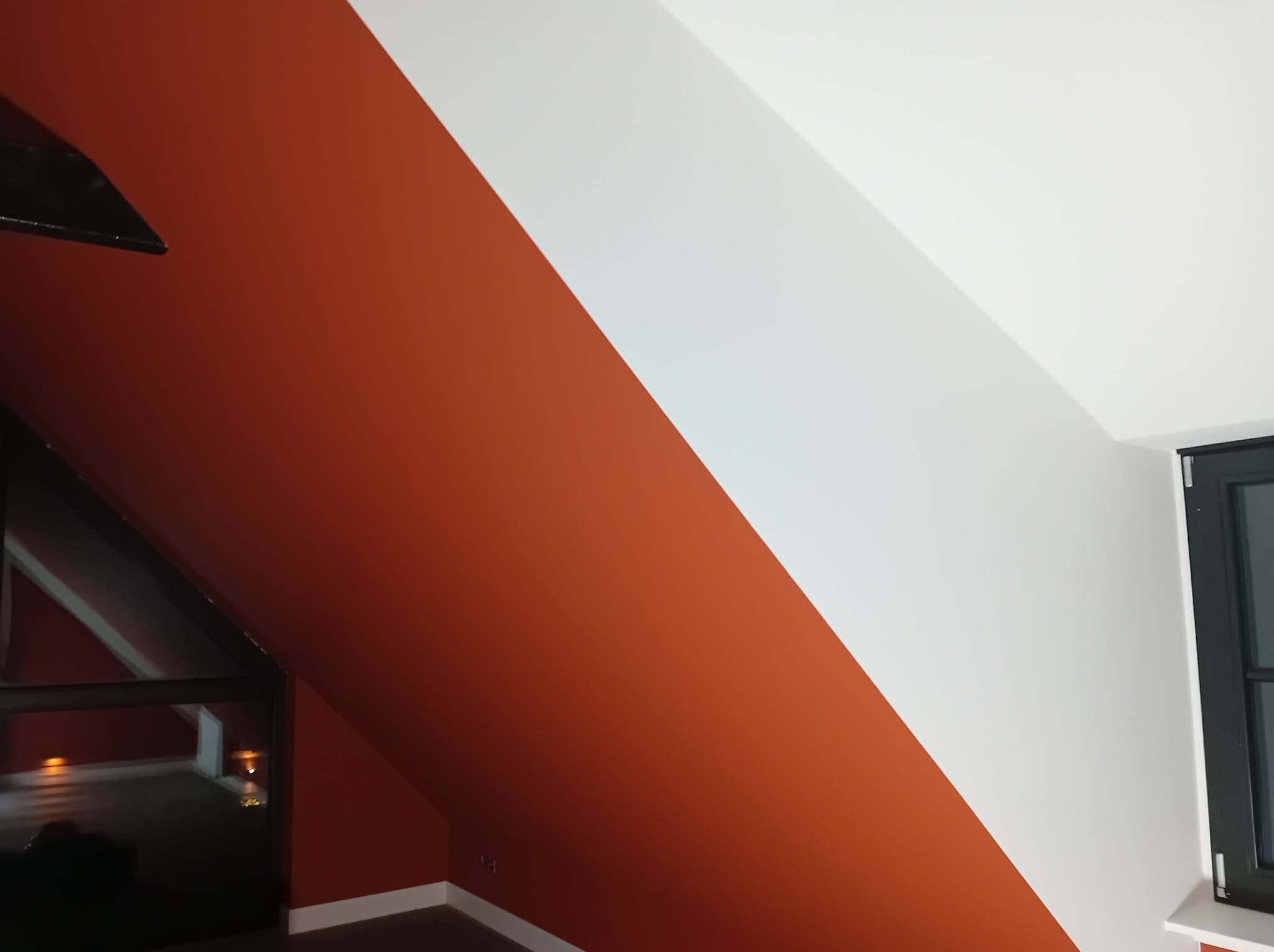 Pomarańczowo-biała ściana na poddaszu, ukośny podział kolorów, widoczne okno i fragment podłogi. Świeżo pomalowane, gładkie powierzchnie.