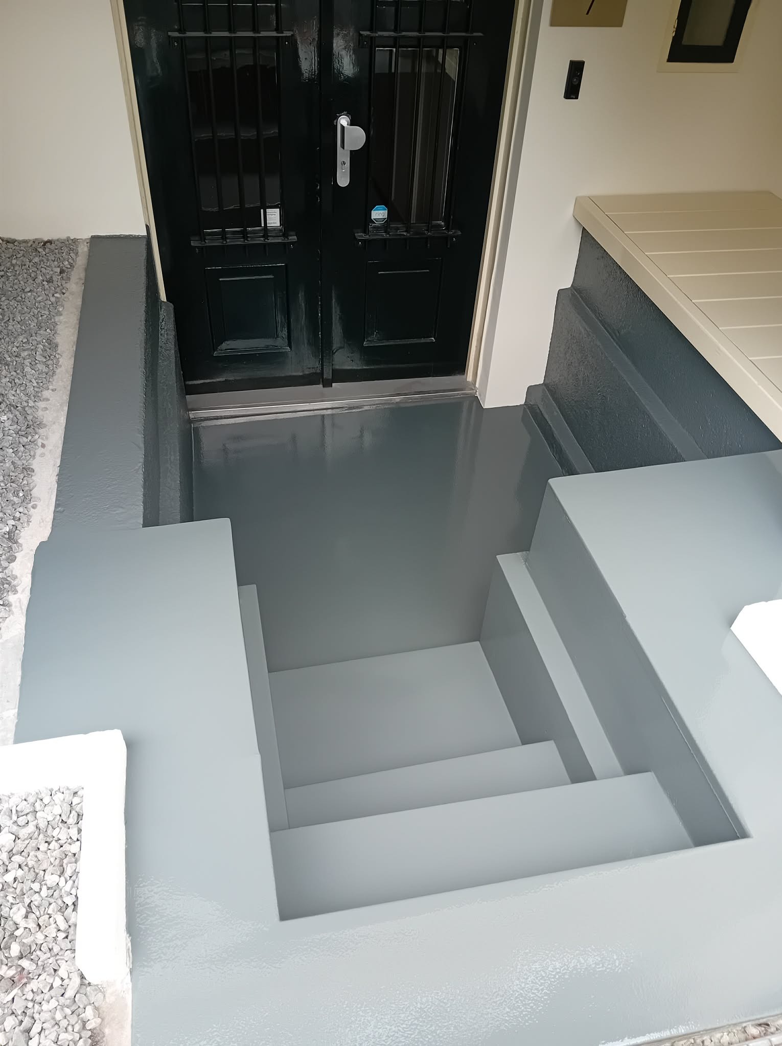 Schody wejściowe po malowaniu na szaro, z widocznymi detalami wykończenia i czarnymi drzwiami wejściowymi. Świeżo pomalowana powierzchnia schodów.