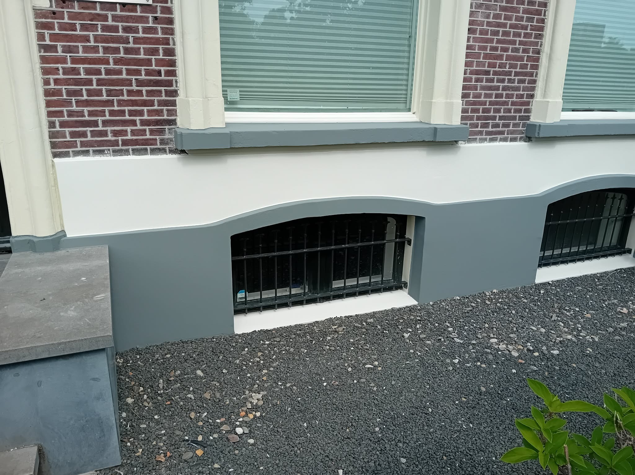 Odnowiona elewacja domu w Łęce Opatowskiej: świeżo pomalowane okna piwniczne z kratami, szara podmurówka i ceglana ściana. Widoczne kamyki dekoracyjne.