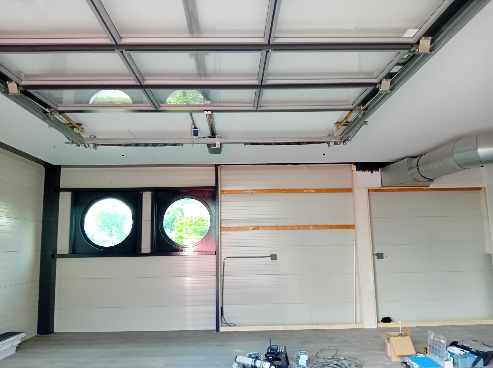 Montaż segmentowej bramy garażowej z oknami, widoczne prowadnice pod sufitem i fragment wentylacji. Surowy stan, narzędzia na podłodze.