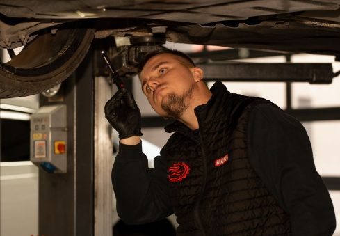 Młody mężczyzna w czarnej kamizelce, z brodą, w warsztacie samochodowym, oświetla lampką podwozie auta na podnośniku, kontrolując stan techniczny pojazdu.