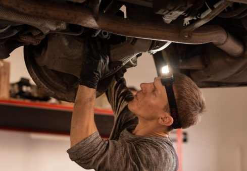 Mechanik z lampą czołową w czarnych rękawicach naprawia podwozie samochodu, skupiając się na elemencie zawieszenia. Widoczne rury wydechowe i elementy podwozia.