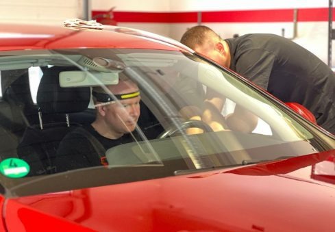 Czerwony samochód w warsztacie: mężczyzna w środku z lampką na głowie, drugi sprawdza szybę. Diagnostyka pojazdu w trakcie naprawy.