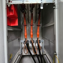 MICHAŁ GRABKA EMG - Wnętrze rozdzielnicy elektrycznej z trzema grubymi kablami w izolacji, widoczne sprężyny i elementy mocujące. Żółty przewód uziemiający po lewej i prawej stronie.