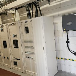 MICHAŁ GRABKA EMG - Rozdzielnia elektryczna z szafami sterowniczymi i przewodami w budynku z białej cegły. Widoczne zabezpieczenia i elementy instalacji elektrycznej.
