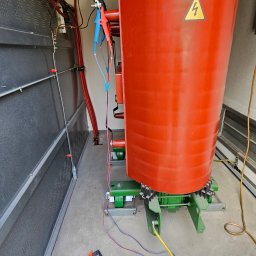 MICHAŁ GRABKA EMG - Transformator w komorze pomiarowej z podłączonym miernikiem. Widoczne kable, oznaczenia ostrzegawcze i elementy konstrukcyjne. Pomiar elektryczny transformatora.