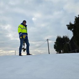 MICHAŁ GRABKA EMG - Elektryk w odblaskowej kurtce, na śniegu, trzyma urządzenie pomiarowe. W tle pochmurne niebo i drzewa. Praca w terenie, zimowa sceneria.