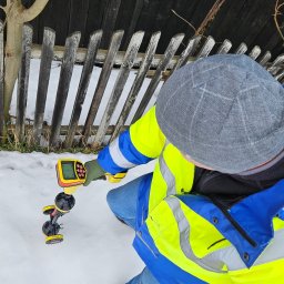 MICHAŁ GRABKA EMG - Elektryk w odblaskowej kurtce używa detektora do pomiarów w zasypanym śniegiem terenie, przy drewnianym płocie i ścianie budynku. Urządzenie RIDGID z wyświetlaczem.