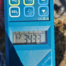 MICHAŁ GRABKA EMG - Miernik Sonel z wyświetlanym wynikiem pomiaru, widoczne przyciski funkcyjne i oznaczenia jednostek pomiarowych. Urządzenie w dłoni, tło z elementami trawy.