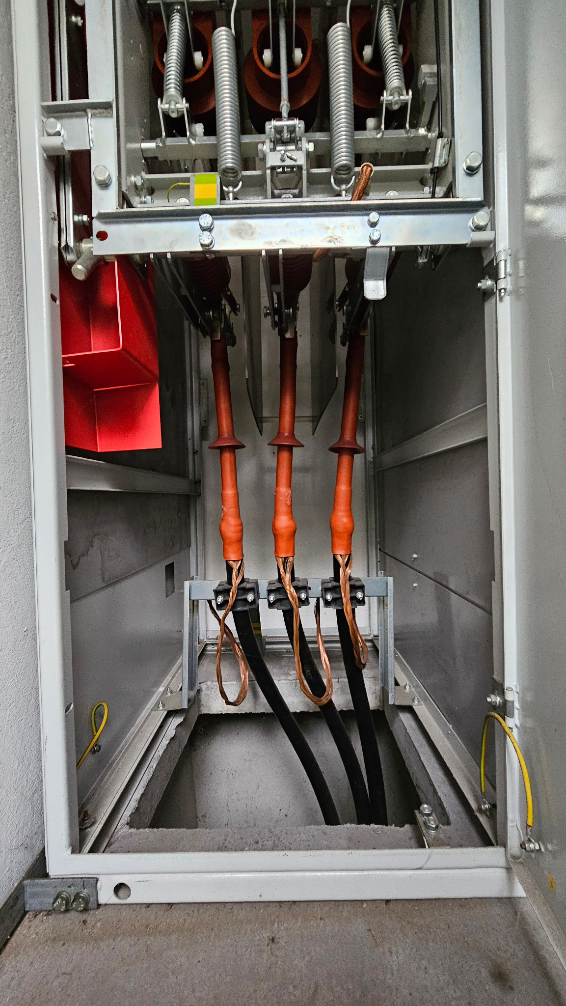 Wnętrze rozdzielnicy elektrycznej z trzema grubymi kablami w izolacji, widoczne sprężyny i elementy mocujące. Żółty przewód uziemiający po lewej i prawej stronie.