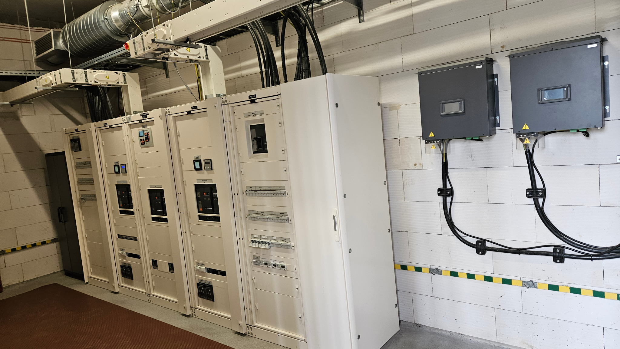 Rozdzielnia elektryczna z szafami sterowniczymi i przewodami w budynku z białej cegły. Widoczne zabezpieczenia i elementy instalacji elektrycznej.