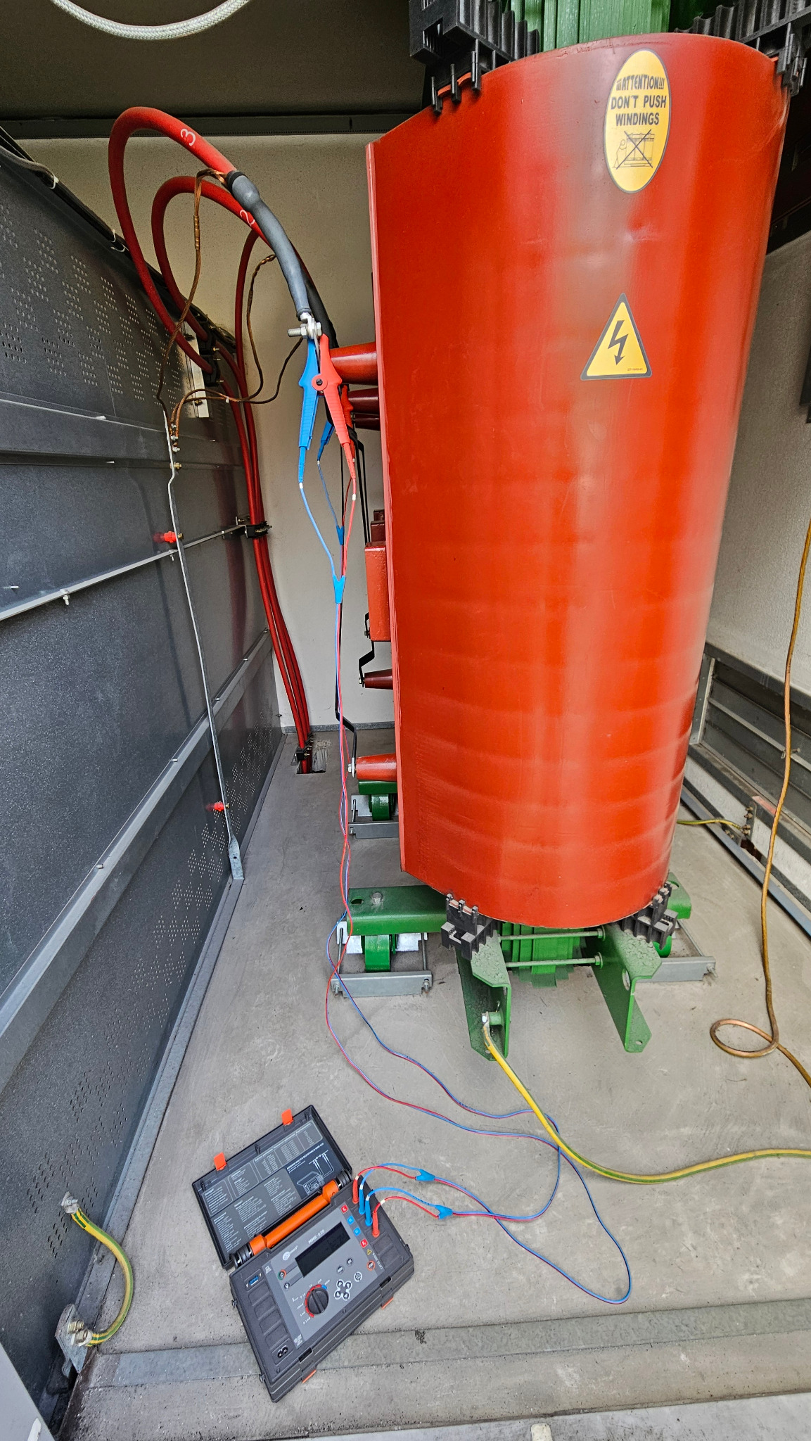 Transformator w komorze pomiarowej z podłączonym miernikiem. Widoczne kable, oznaczenia ostrzegawcze i elementy konstrukcyjne. Pomiar elektryczny transformatora.