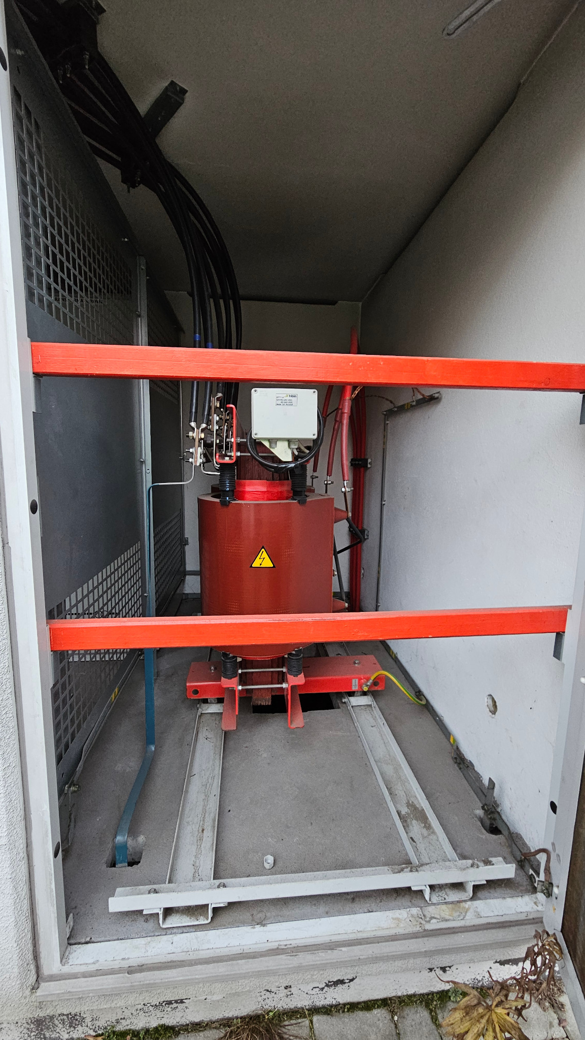Czerwony transformator z symbolem ostrzegawczym wewnątrz szarej szafy elektrycznej z widocznymi kablami i elementami instalacji. Ujęcie z bliska, oświetlenie naturalne.