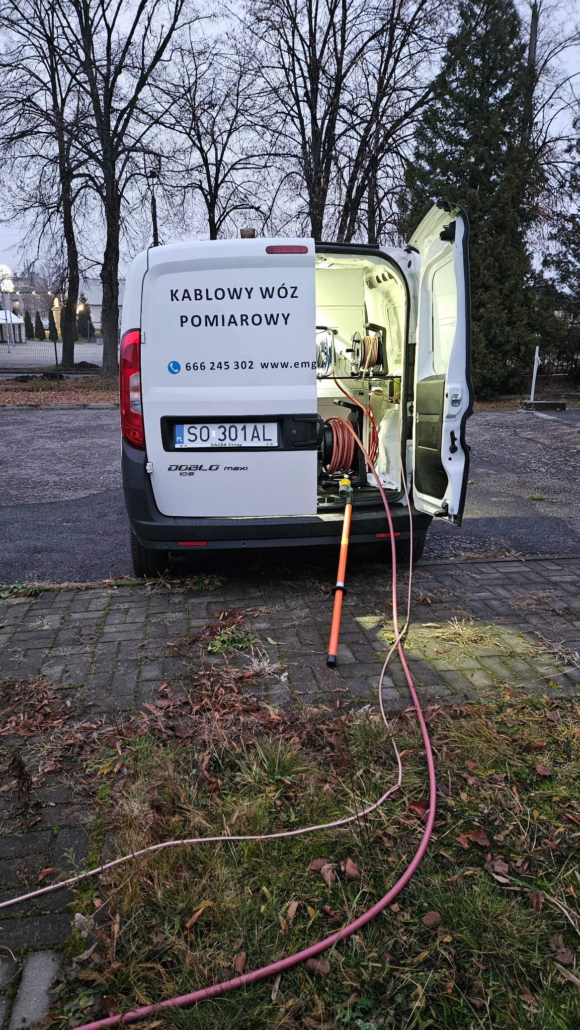 Otwarty tył białego samochodu dostawczego z napisem 'Kablowy Wóz Pomiarowy', widoczne narzędzia pomiarowe i przewody. Tło stanowią drzewa i budynki.