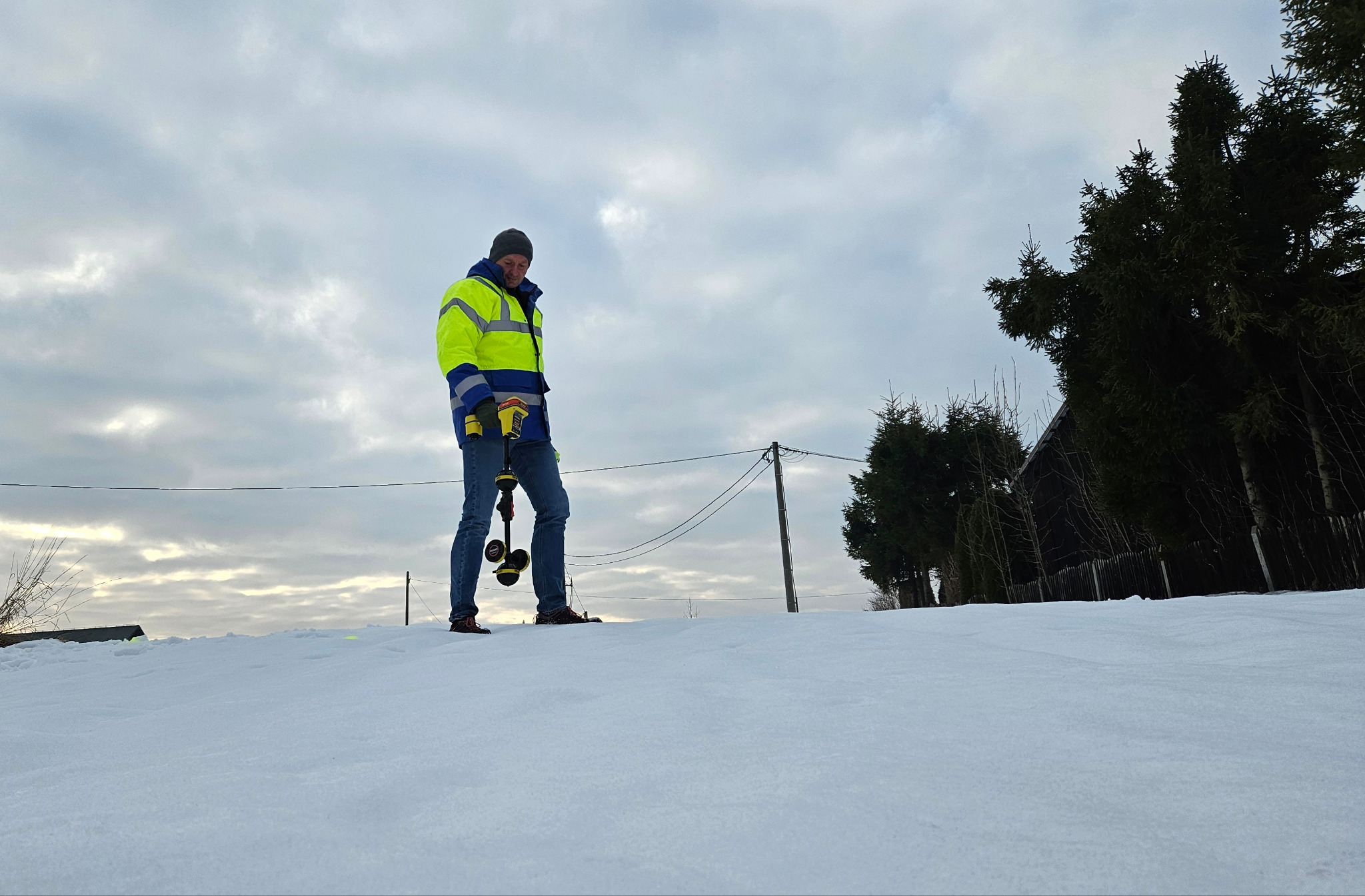 Elektryk w odblaskowej kurtce, na śniegu, trzyma urządzenie pomiarowe. W tle pochmurne niebo i drzewa. Praca w terenie, zimowa sceneria.