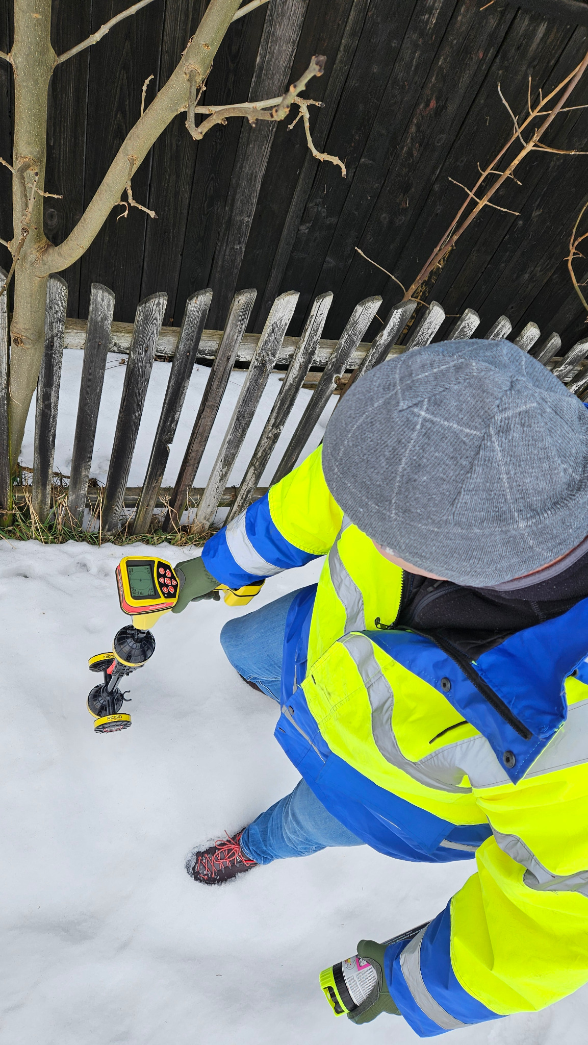 Elektryk w odblaskowej kurtce używa detektora do pomiarów w zasypanym śniegiem terenie, przy drewnianym płocie i ścianie budynku. Urządzenie RIDGID z wyświetlaczem.