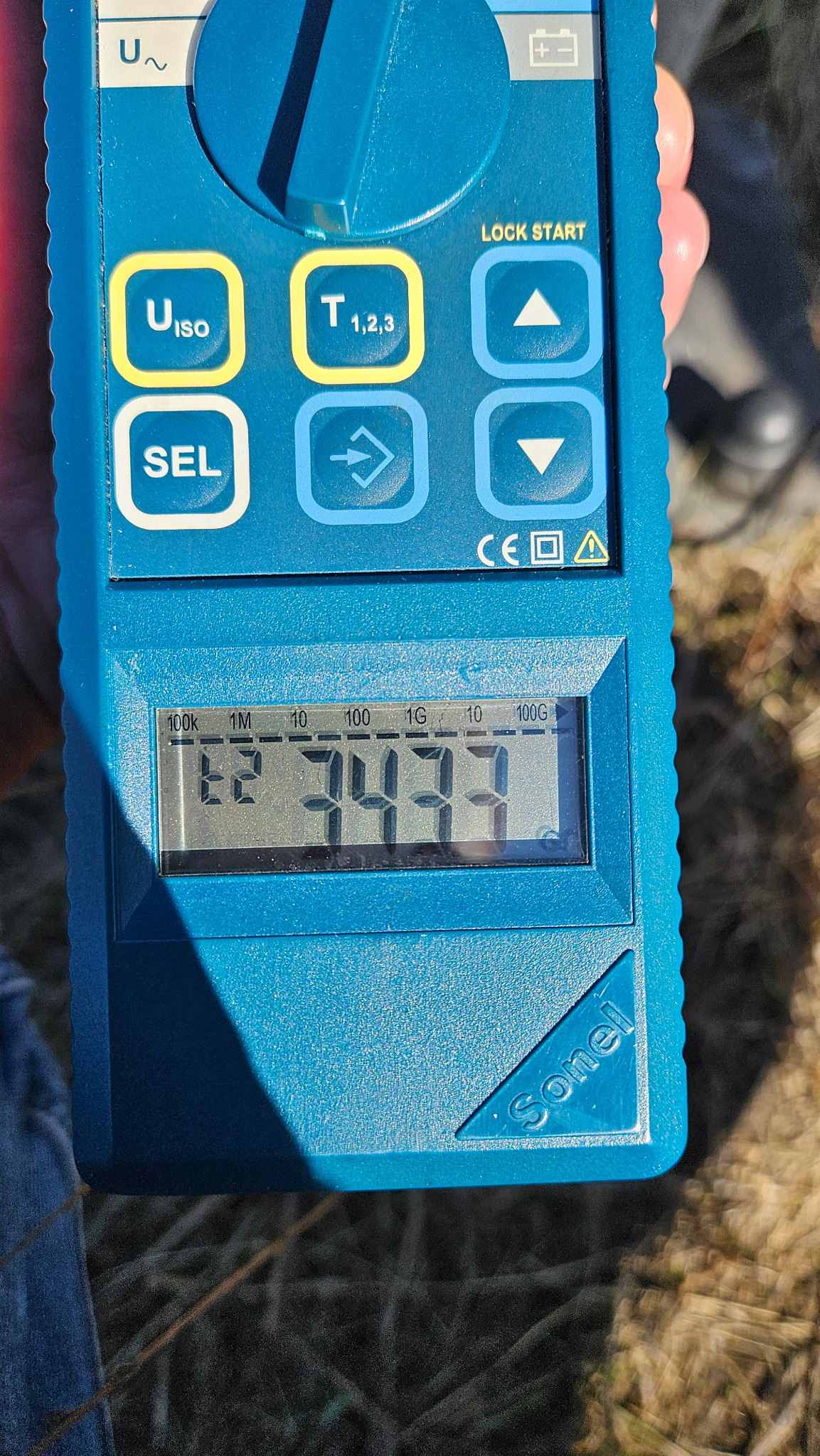 Miernik Sonel z wyświetlanym wynikiem pomiaru, widoczne przyciski funkcyjne i oznaczenia jednostek pomiarowych. Urządzenie w dłoni, tło z elementami trawy.