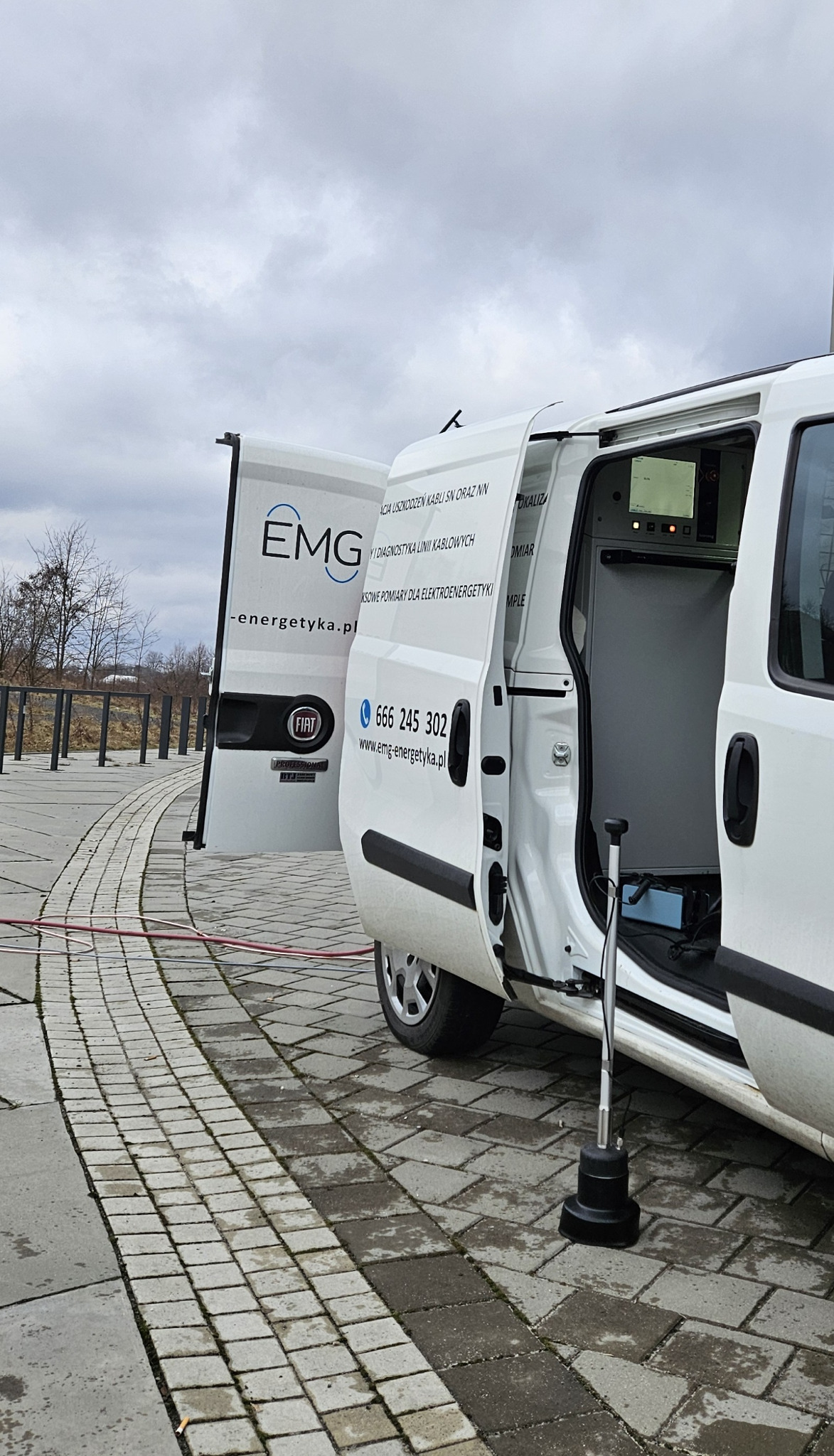 Biały samochód EMG-energetyka z otwartymi drzwiami, sprzęt pomiarowy w środku, kabel na zewnątrz. Diagnostyka i lokalizacja uszkodzeń kabli SN i NN.