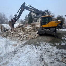 Przedsiębiorstwo Wielobranżowe Multitrans Mateusz Sieprawski - Żółta koparka Volvo na tle gruzów i częściowo rozebranego budynku w zimowej scenerii. Widoczny dom w tle.