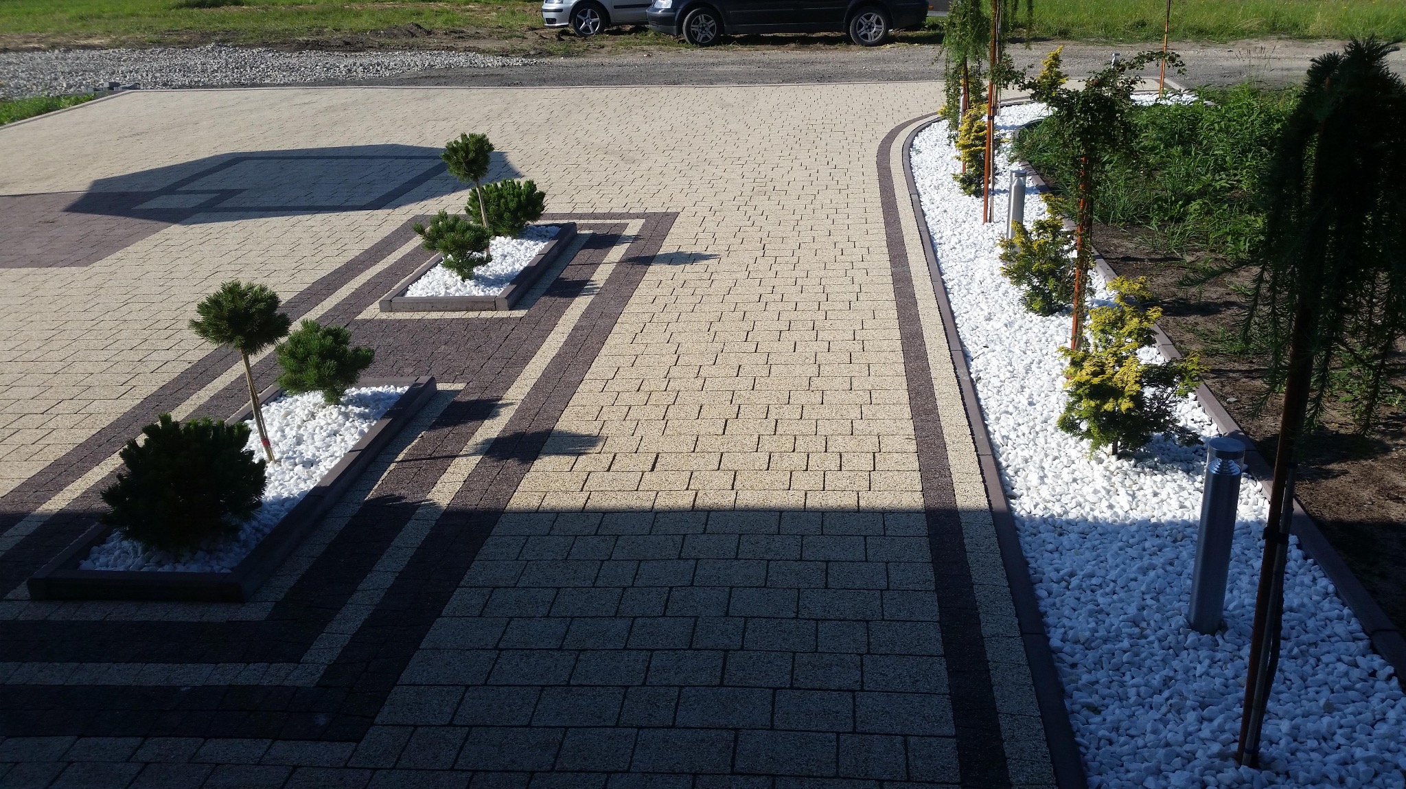 Estetyczna nawierzchnia z kostki brukowej w odcieniach beżu i brązu, z dekoracyjnymi rabatami wypełnionymi białym kamieniem i niskimi drzewkami. Widok z góry, samochody w tle.