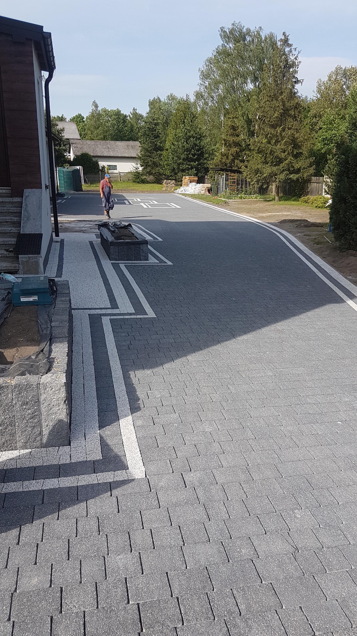 Ułożona szara kostka brukowa z białymi pasami, tworząca wzór. W tle budynki i osoba. Widoczne elementy wykończeniowe i narzędzia.