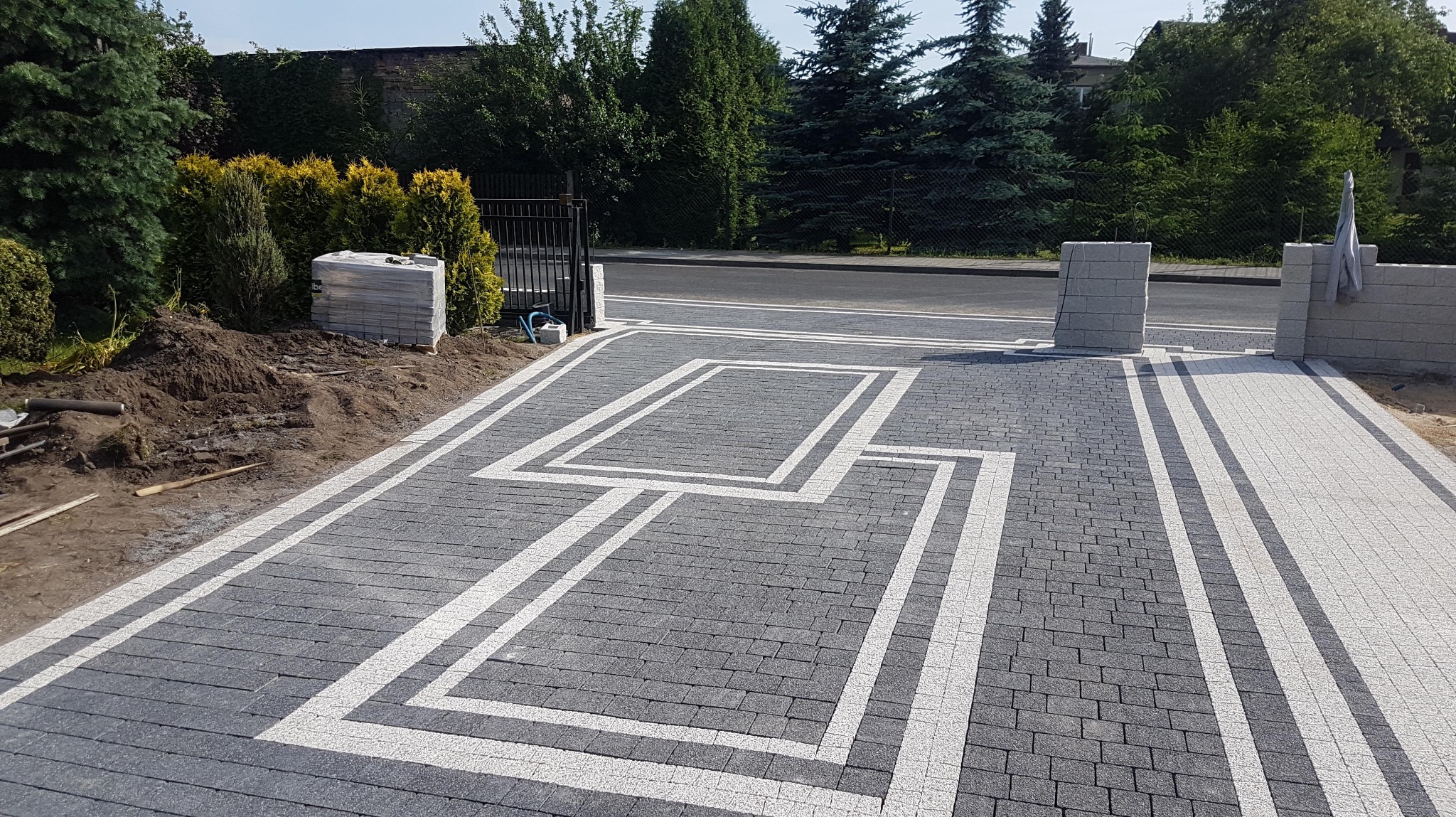 Ukończony podjazd z kostki brukowej w odcieniach szarości i bieli, z geometrycznym wzorem. Widoczne elementy ogrodzenia i przygotowane kostki do dalszej pracy.