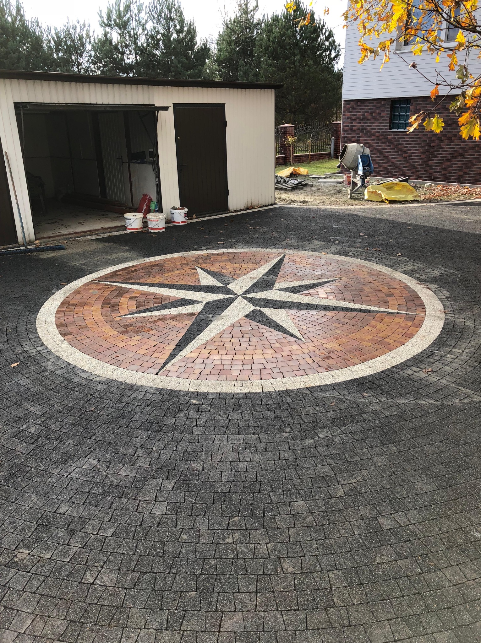 Okrągły podjazd z kostki brukowej w odcieniach szarości, brązu i bieli, ułożony we wzór kompasu. W tle garaż, dom i betoniarka.