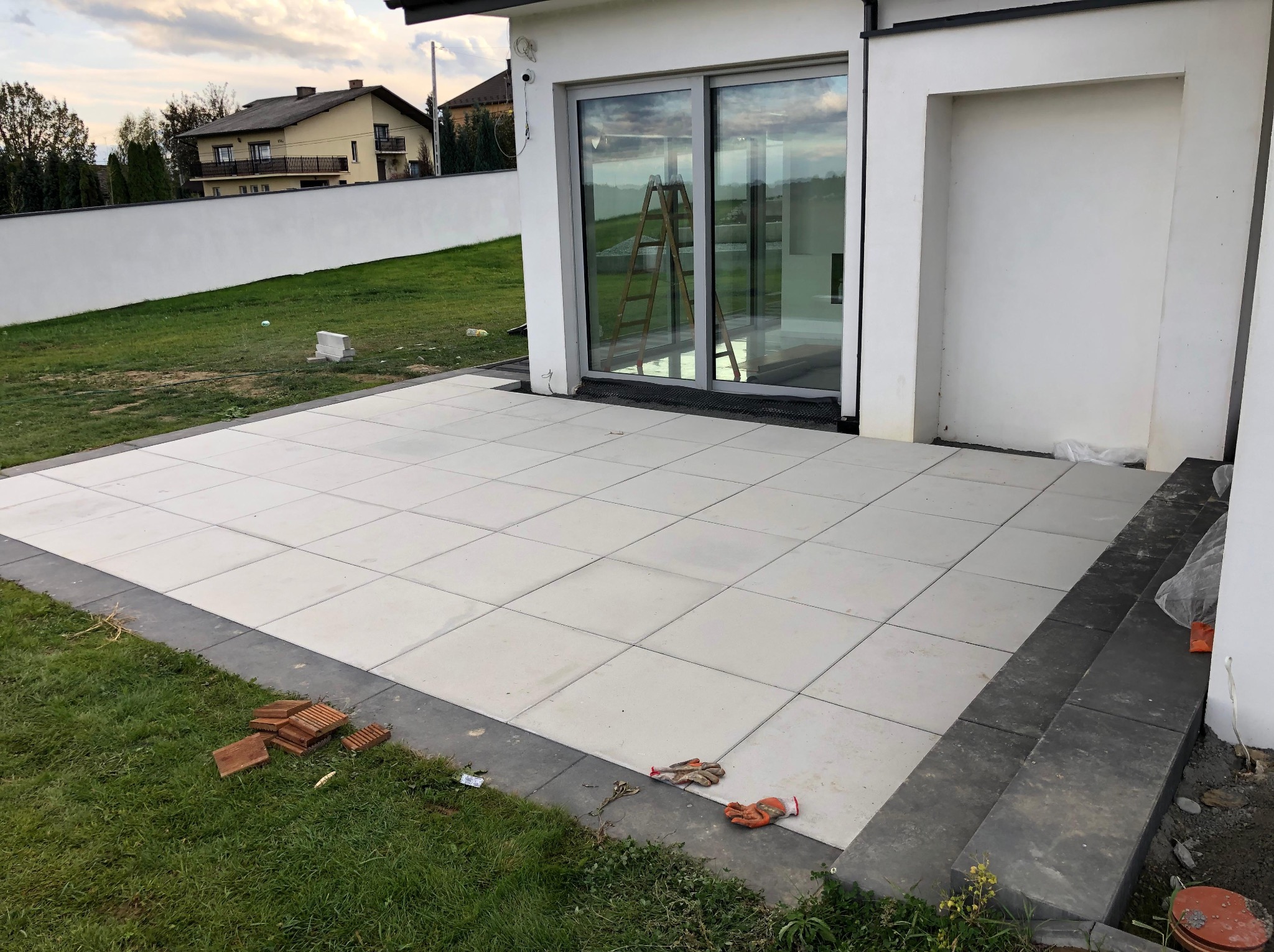 Nowoczesny taras z jasnych płyt, obrzeża z ciemnej kostki. Widoczne rękawice robocze i fragmenty niezakończonych prac. Tło: biały budynek i trawnik.