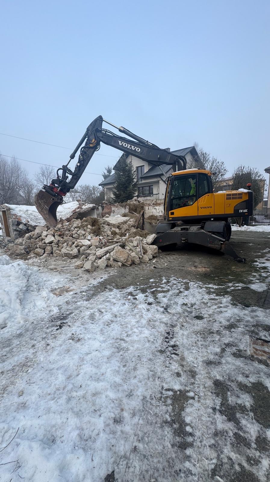 Żółta koparka Volvo na tle gruzów i częściowo rozebranego budynku w zimowej scenerii. Widoczny dom w tle.