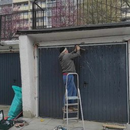 Henryk Rolka - Mężczyzna na drabinie montuje bramę garażową, narzędzia obok. W tle blok mieszkalny i kwitnące drzewa. Ujęcie z dołu, podkreślające etap prac.