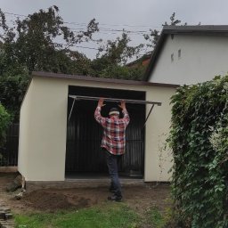 Henryk Rolka - Montaż bramy garażowej uchylnej w blaszanym garażu. Mężczyzna w koszuli w kratę podtrzymuje otwartą bramę. Widok z zewnątrz, pochmurne niebo.