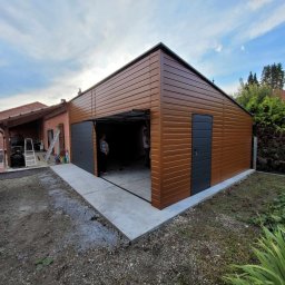 Henryk Rolka - Brązowy garaż blaszany z szarą bramą segmentową i drzwiami wejściowymi, widok z zewnątrz na betonowy podjazd i częściowo otwartą bramę. Budynek w otoczeniu zieleni.