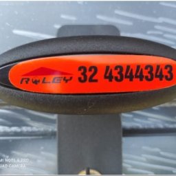 Henryk Rolka - Detal bramy garażowej z numerem kontaktowym na pomarańczowej naklejce. Widoczny zamek i fragment konstrukcji bramy.