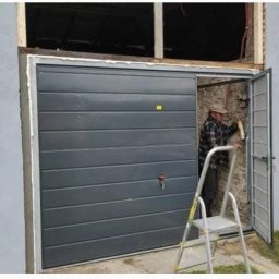 Henryk Rolka - Montaż szarej bramy garażowej segmentowej. Mężczyzna na drabinie, w tle budynek z otwartymi drzwiami i oknem. Widoczne narzędzia i fragment samochodu.