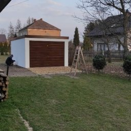 Henryk Rolka - Murowany garaż z brązową bramą segmentową, świeżo pomalowany na biało. Osoba maluje ścianę. Na podjeździe żwir. W tle domy i drzewa.