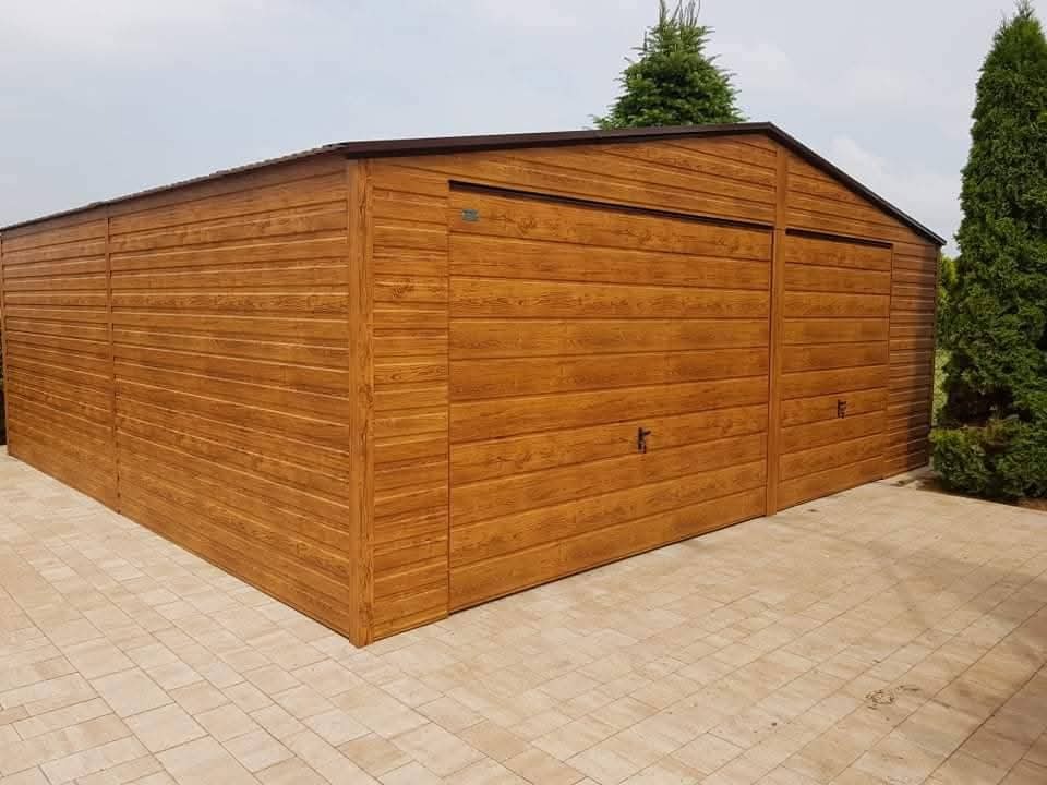 Blaszany garaż dwustanowiskowy z bramami segmentowymi w kolorze drewna, na kostce brukowej. Widok z boku, z zielonymi drzewami w tle.