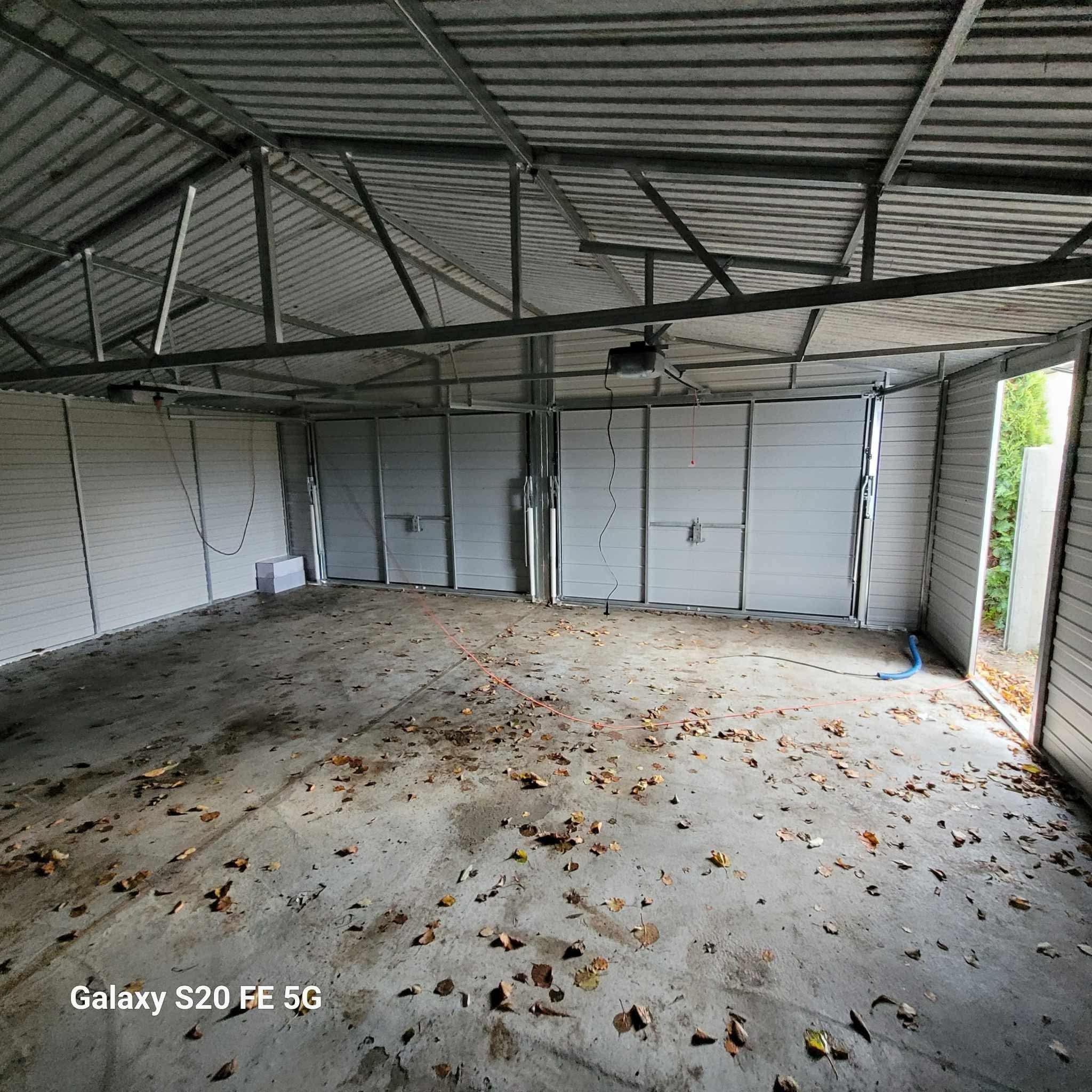 Wnętrze garażu blaszanego z otwartymi drzwiami i widoczną konstrukcją dachu. Na podłodze leżą liście. Widoczny automat do bramy.