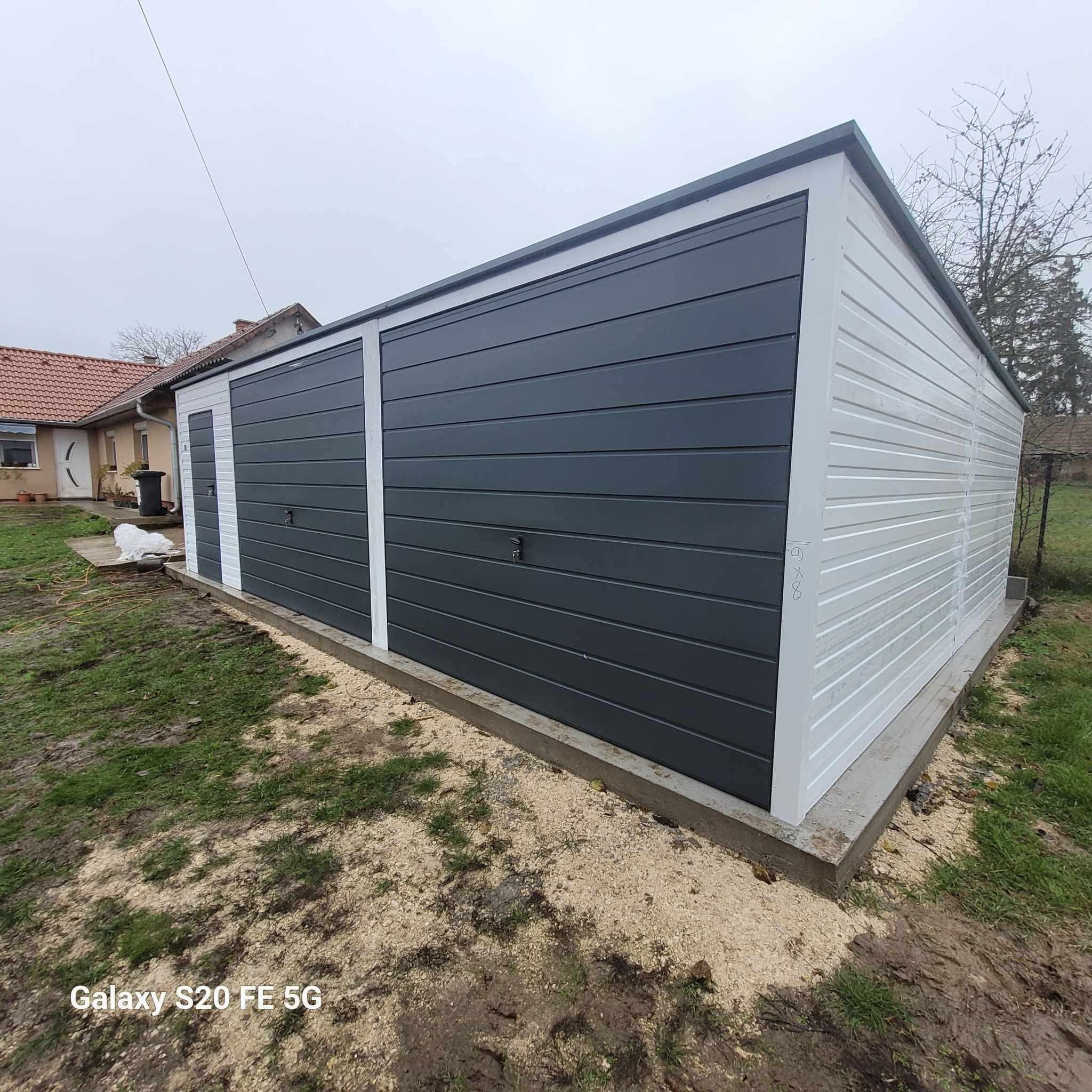 Nowoczesny garaż blaszany dwustanowiskowy, z szarą bramą segmentową i białymi ścianami bocznymi, na betonowej wylewce, z widocznym domem w tle.