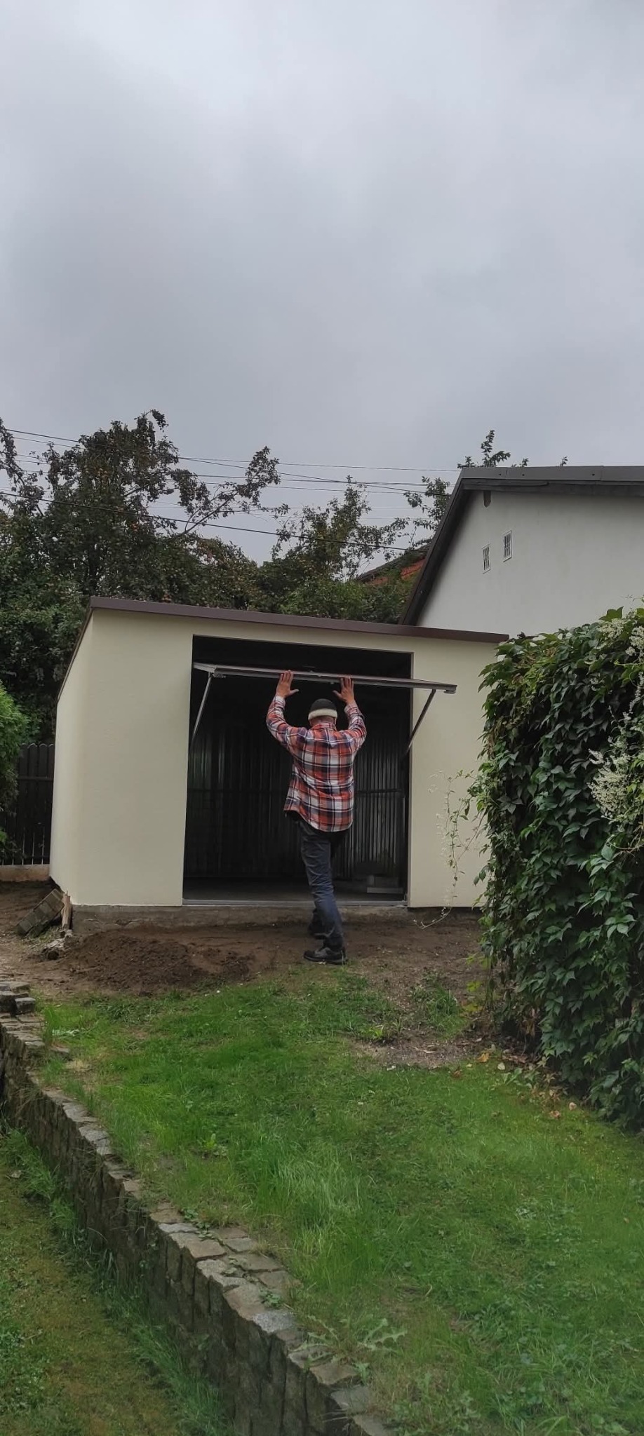 Montaż bramy garażowej uchylnej w blaszanym garażu. Mężczyzna w koszuli w kratę podtrzymuje otwartą bramę. Widok z zewnątrz, pochmurne niebo.