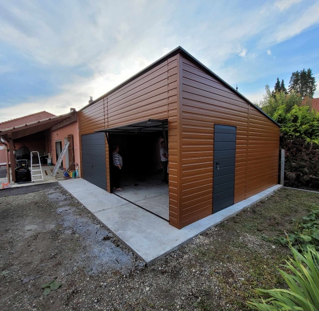 Brązowy garaż blaszany z szarą bramą segmentową i drzwiami wejściowymi, widok z zewnątrz na betonowy podjazd i częściowo otwartą bramę. Budynek w otoczeniu zieleni.