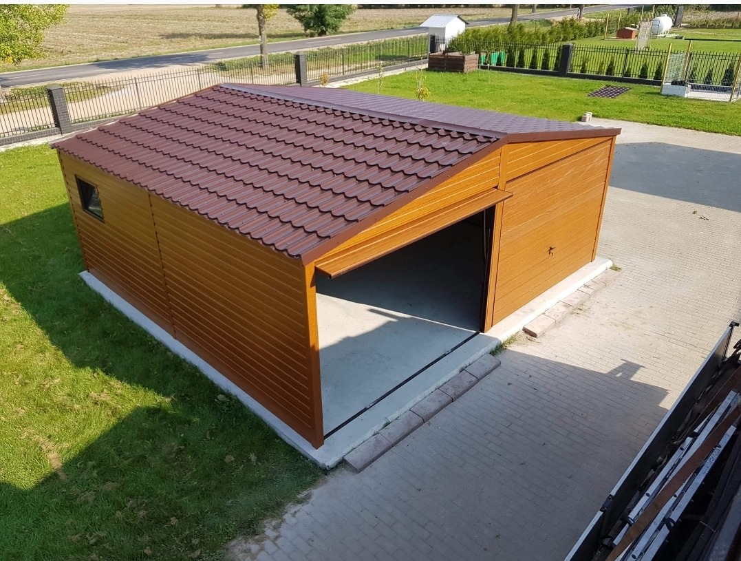 Garaż blaszany z brązowym dachem i drewnopodobną elewacją, częściowo otwarty, widok z góry na tle zielonego trawnika i ogrodzenia.