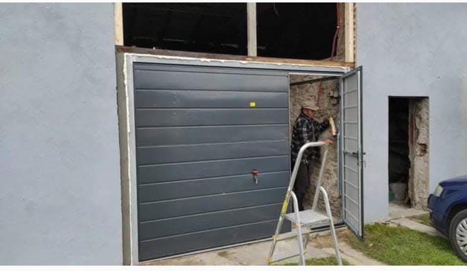 Montaż szarej bramy garażowej segmentowej. Mężczyzna na drabinie, w tle budynek z otwartymi drzwiami i oknem. Widoczne narzędzia i fragment samochodu.