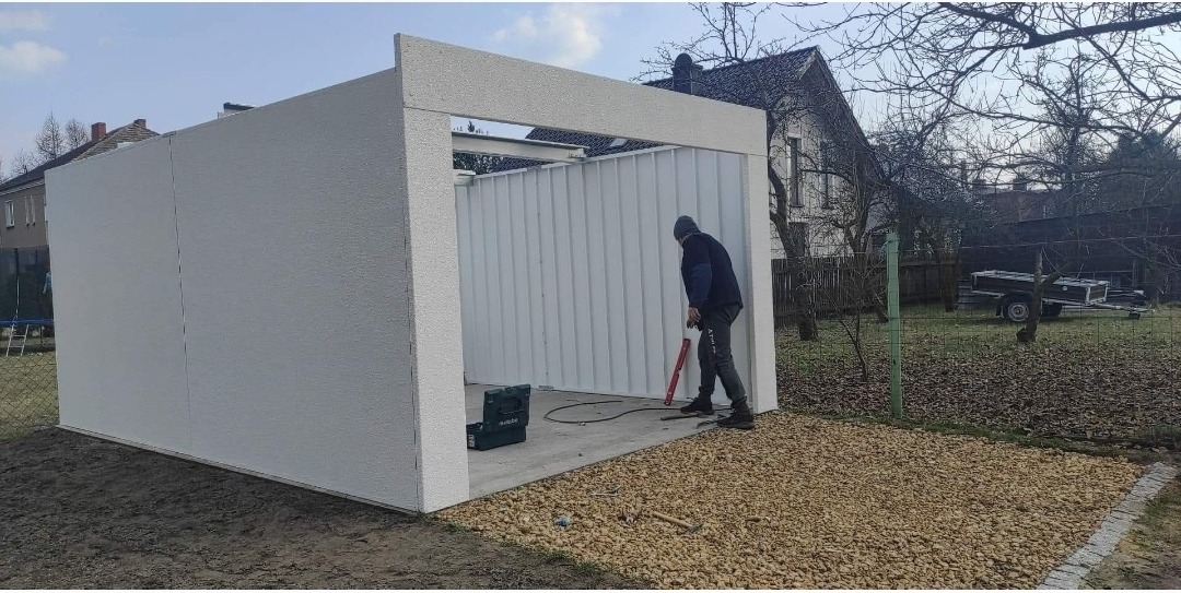 Montaż garażu blaszanego: pracownik z poziomicą przy konstrukcji z białych paneli, widoczny sprzęt i podłoże z kamieni. W tle dom i przyczepa.