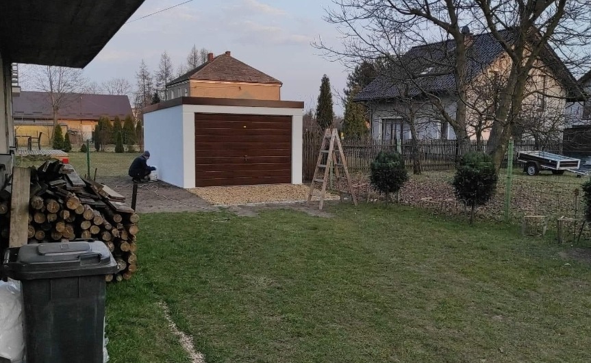 Murowany garaż z brązową bramą segmentową, świeżo pomalowany na biało. Osoba maluje ścianę. Na podjeździe żwir. W tle domy i drzewa.